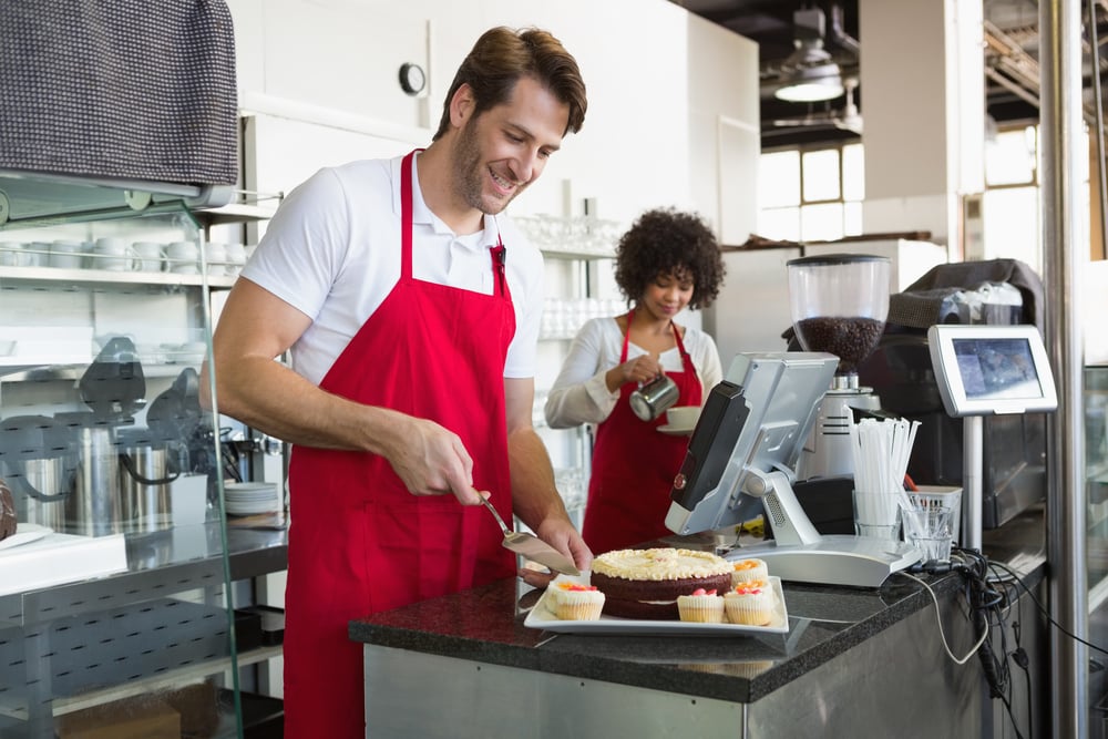 Smiling waiter slicing cake with waitress behind him at the bakery